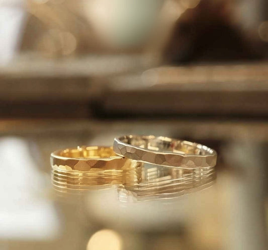 Two elegant, textured rings—one gold, one silver—rest side by side on a reflective surface, softly lit to highlight their craftsmanship and shine.
