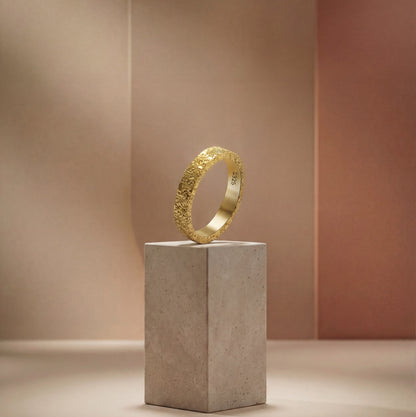 A textured gold ring labeled "925" stands on a stone pedestal against a soft gradient beige and peach background, conveying elegance and simplicity.