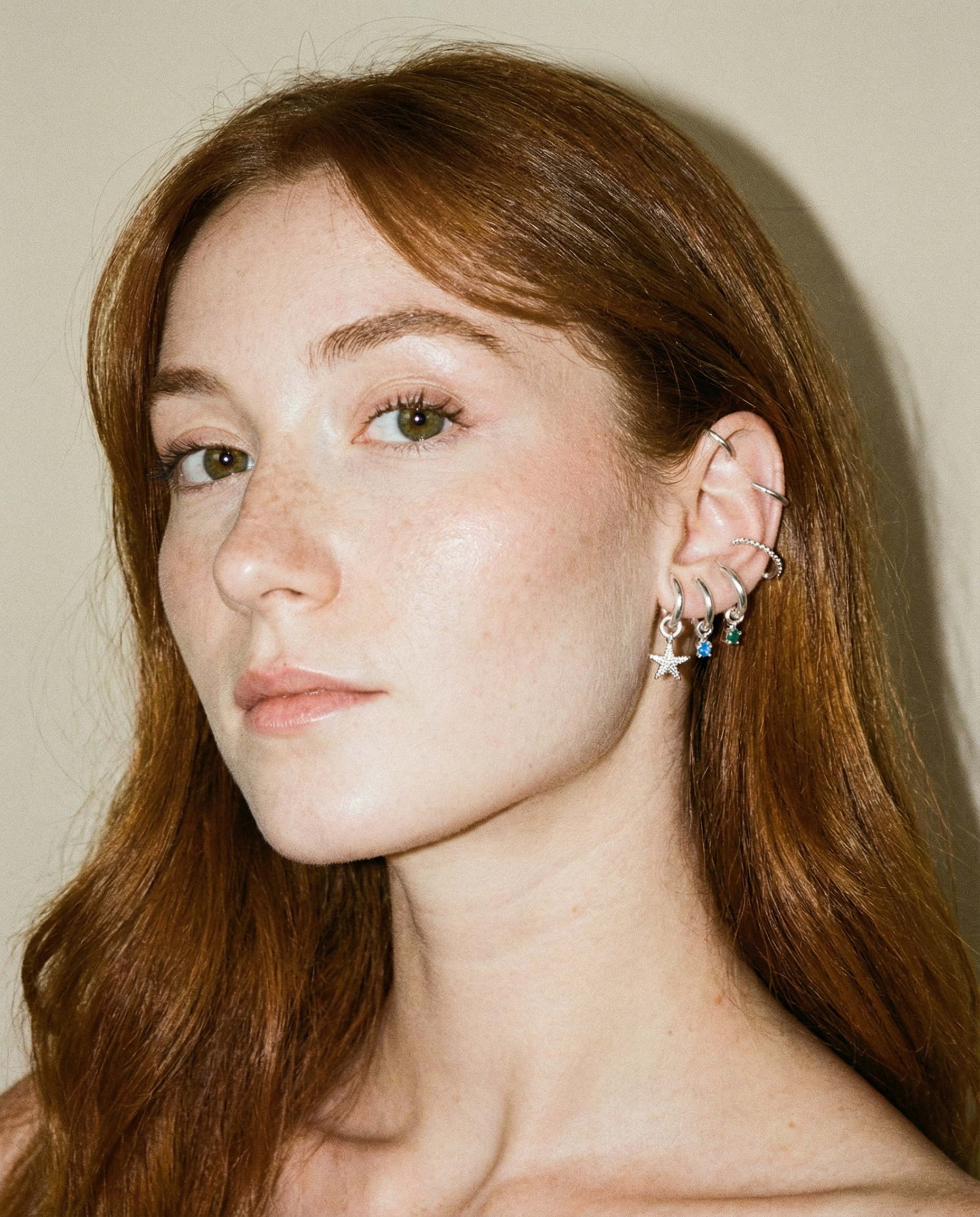 A woman with long red hair and fair skin looks at the camera. She wears multiple silver earrings with blue accents on her left ear against a plain light background.