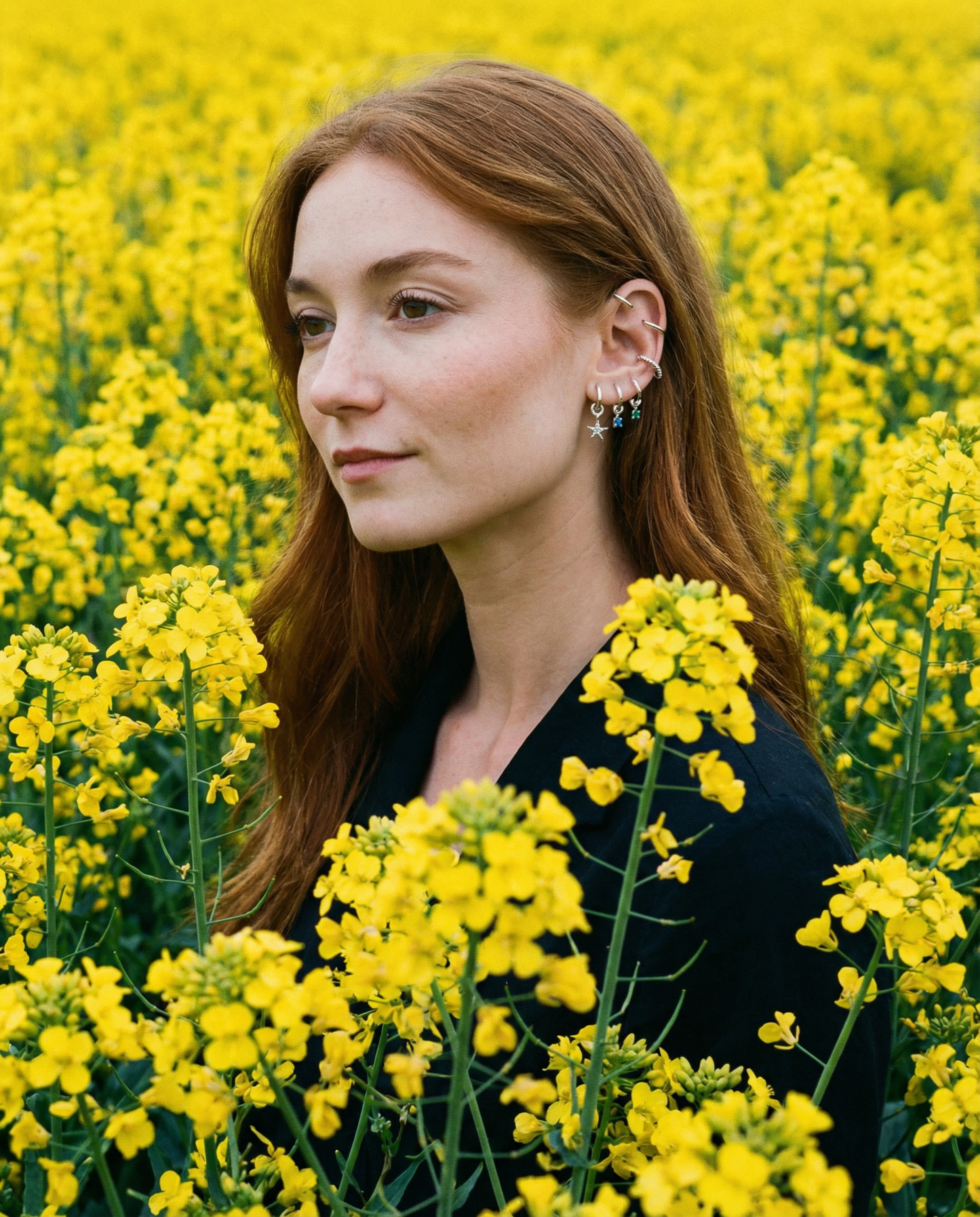 A woman with long red hair and multiple earrings stands in a field of yellow flowers, wearing a black top.