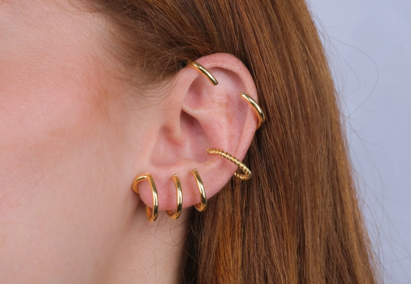 A close-up of a person's ear adorned with multiple gold hoop earrings along the cartilage and lobe, set against light brown hair. The style conveys a modern and edgy fashion statement.