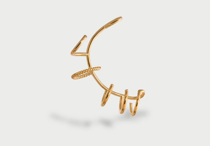 A modern, abstract gold ear cuff with a spiraling, open design, featuring a textured band. It casts a soft shadow on a light gray background.