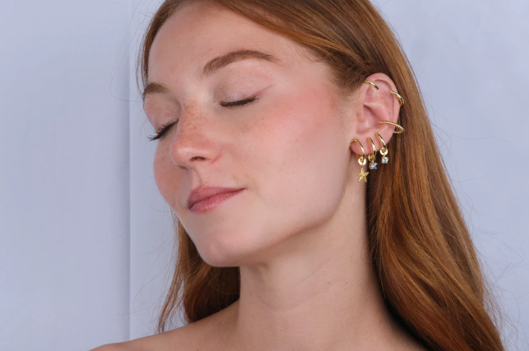 Young woman with closed eyes smiles subtly, showcasing multiple gold earrings on her ear, including hoops and dangling stars, against a light background.